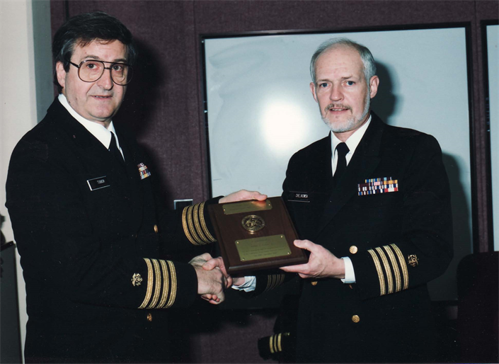 CAPT Chelikowsky receiving a plaque from CAPT Touch, both in service dress blue uniforms.