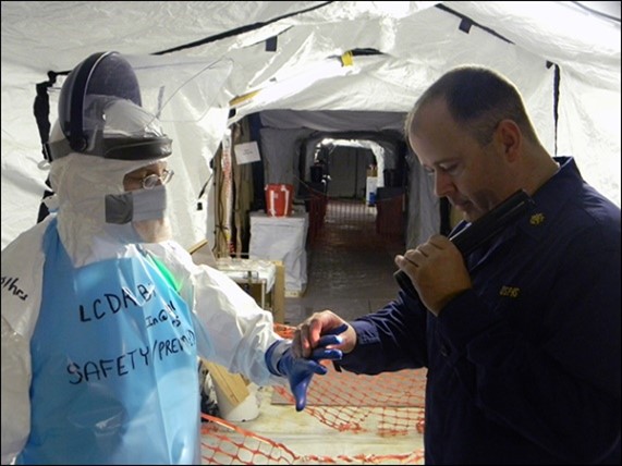 LCDR Adams in uniform inspects another officer wearing full PPE in the medical unit